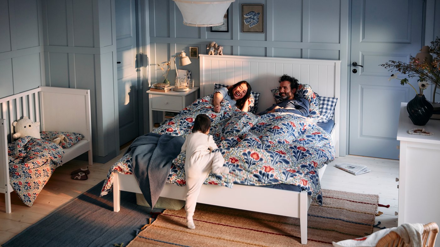 Une chambre à coucher où un couple est allongé dans le lit, tandis qu'un tout-petit grimpe pour les rejoindre.
