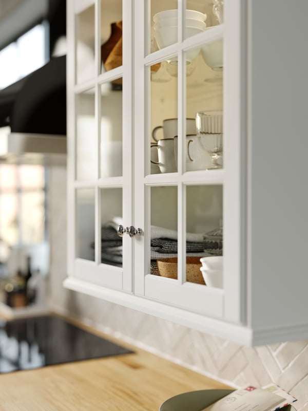 A cabinet with BODBYN glass doors in off-white with shelves holding GLADELIG mugs in grey and VARDAGEN bowls in off-white.
