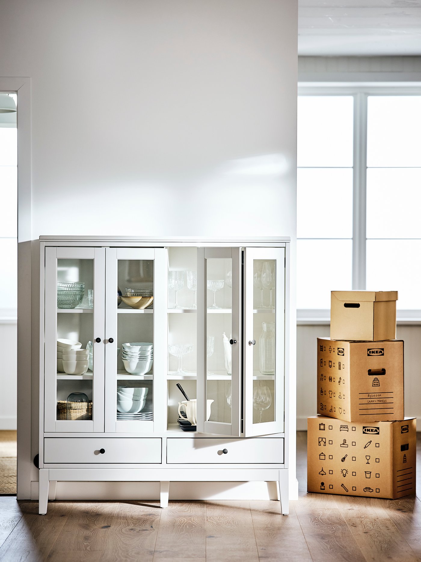 Ein Raum mit weissen Wänden und Böden ist bis auf eine IDANÄS Vitrine mit halb klappbaren Türen und ein paar Umzugskartons komplett leer.