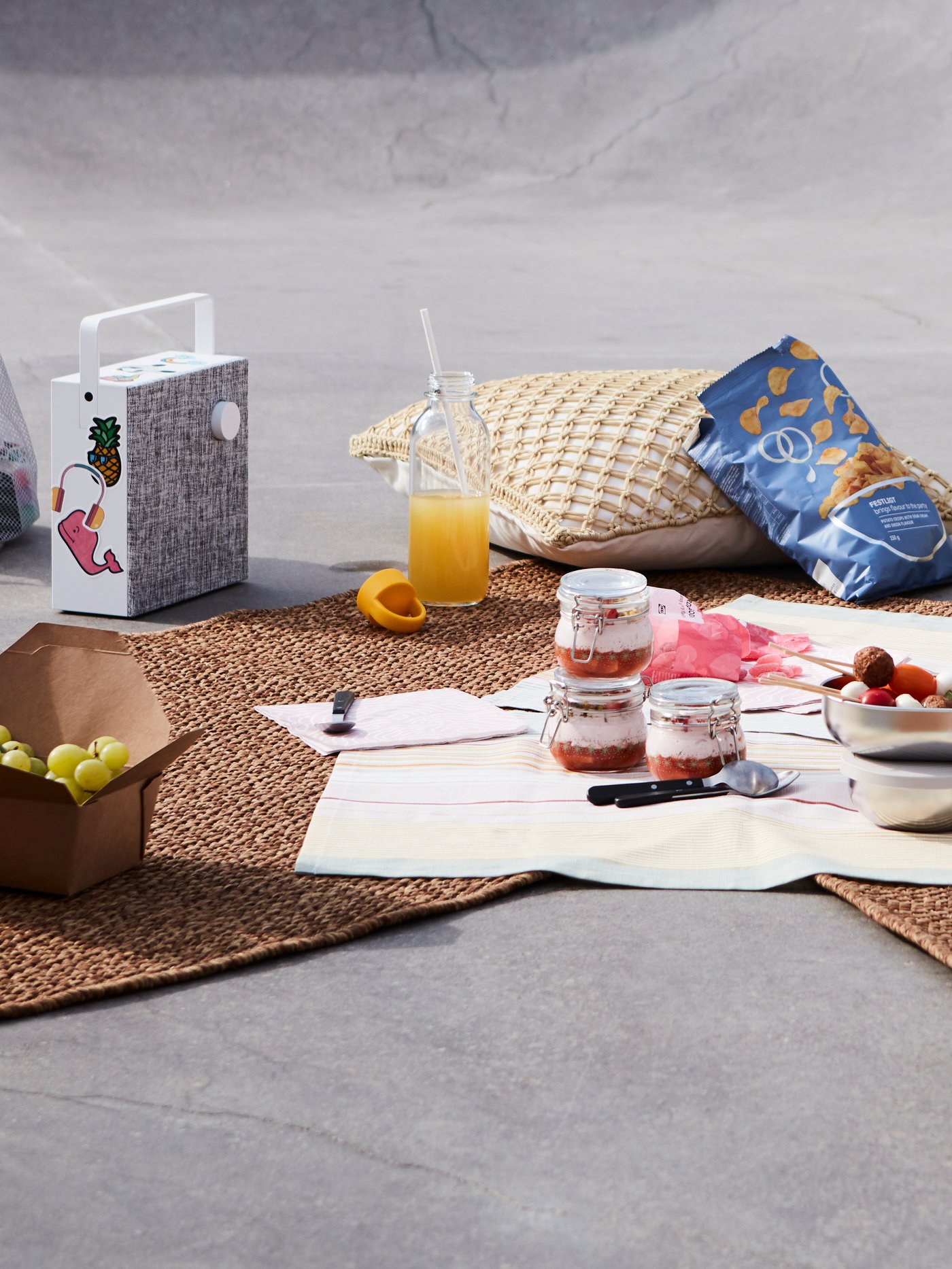 Et picnicarrangement med desserter i glaskrukker, juice, chips, et udendørs tæppe og puder i et bymiljø.