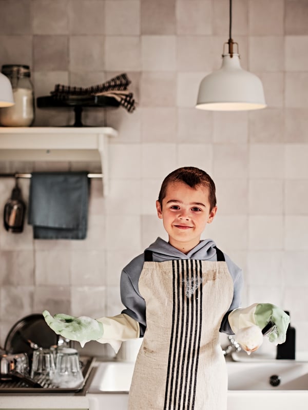 A child in a kitchen in front of a sink wearing a MARIATHERES apron and soapy RINNIG cleaning gloves.
