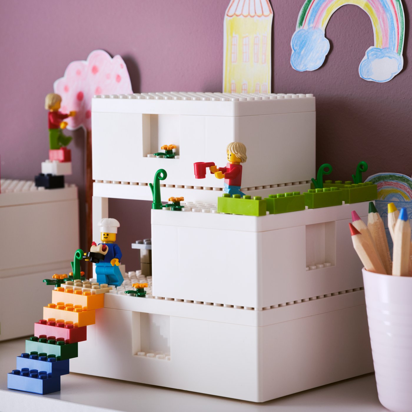 3 cajas BYGGLEK blancas montadas una sobre otra. En sus superficies hay figuras de LEGO montadas armando un jardín con plantas y flores.
