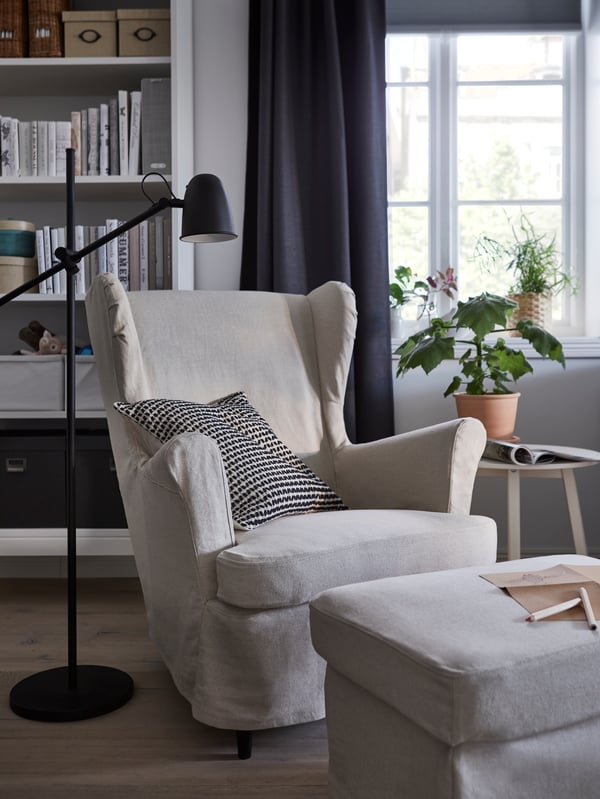 A STRANDMON armchair and footstool with off-white covers, in front of a window and a bookcase, with a black floor lamp by it.