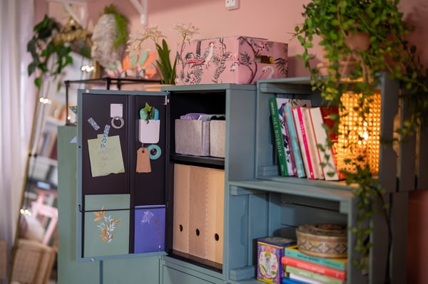 Een groene IKEA kast met vakken gevuld met boeken, dozen, planten en decoratieve objecten in een kleurrijke kamer