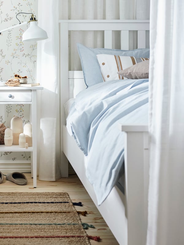 Una lámpara de trabajo RANARP sobre una mesita de noche junto a una cama HEMNES manchada de blanco vestida con ropa de cama BERGPALM de rayas azules.
