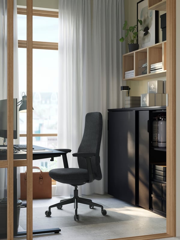 Dans une petite pièce baignée de lumière, poste de travail avec chaise GRÖNFJÄLL noire et rangement à portes coulissantes GALANT noir.