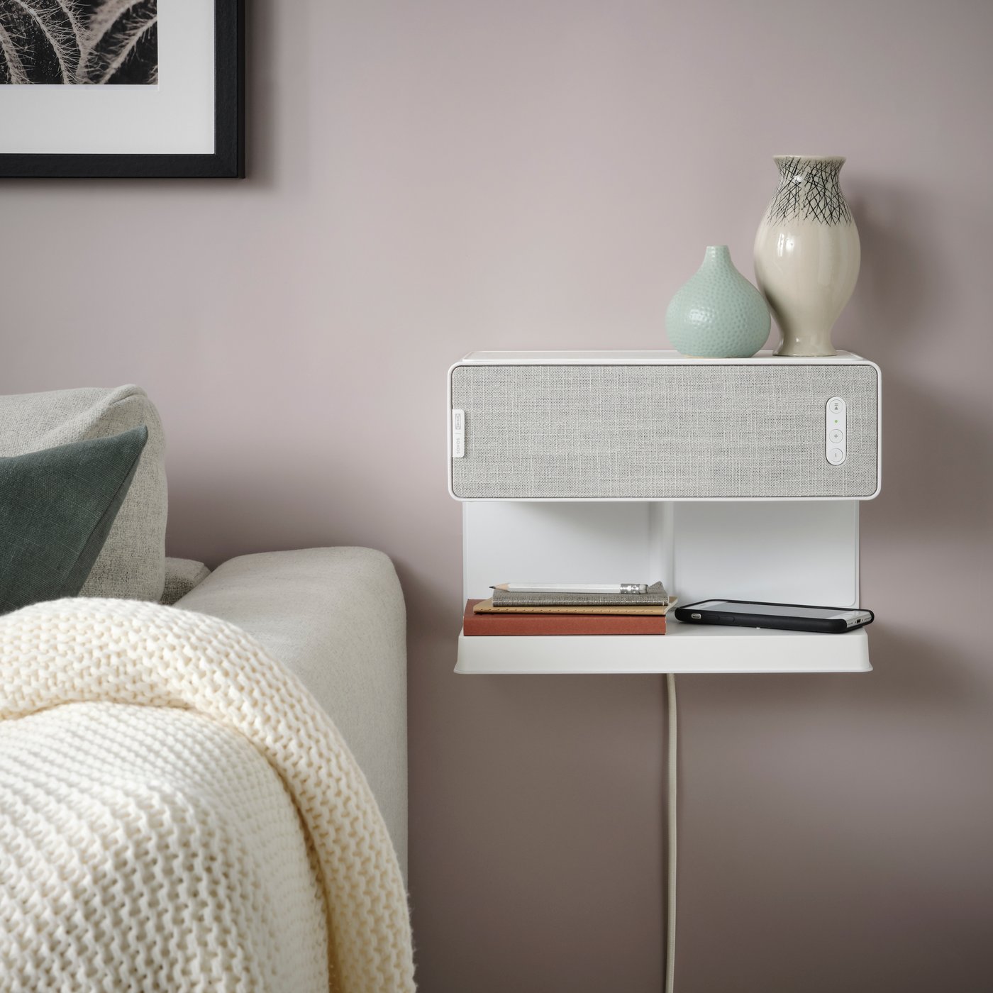 A white SYMFONISK Wi-Fi bookshelf speaker is hanging on the wall in a living room next to a white sofa.