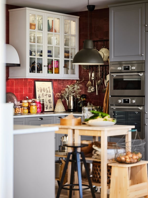 A pair of BEKVÄM birch trolleys, produce on top and accessories on their shelves, stand in the middle of a red-tiled kitchen.