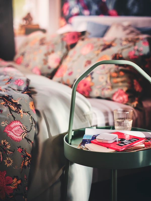 A white STYRBAR remote control placed on a BURVIK side table next to a bed dressed in a NÄSSELKLOCKA duvet set.