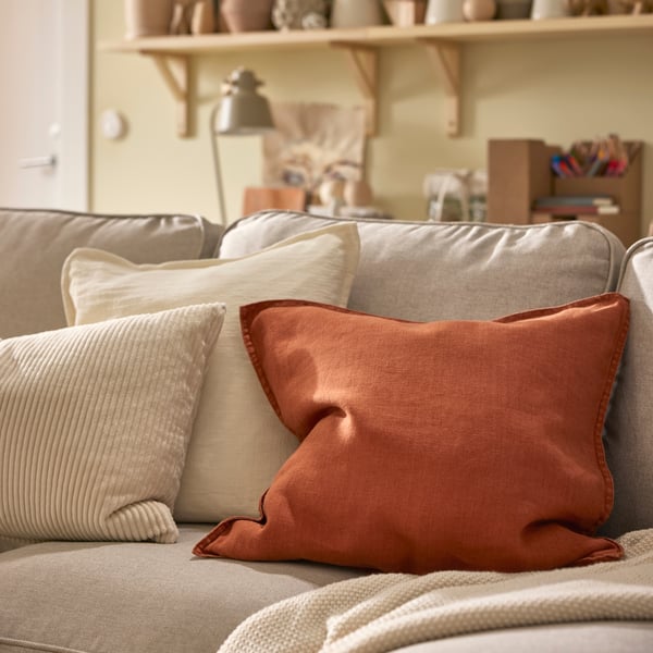 A throw and three cushions, including one with a red-brown DYTÅG cushion cover, lie on a light beige EKTORP sofa.