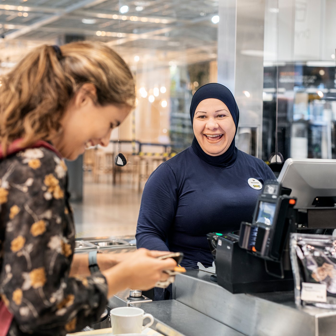 Een IKEA kassamedewerker in een hidjab en een klant lachen aan de kassa.