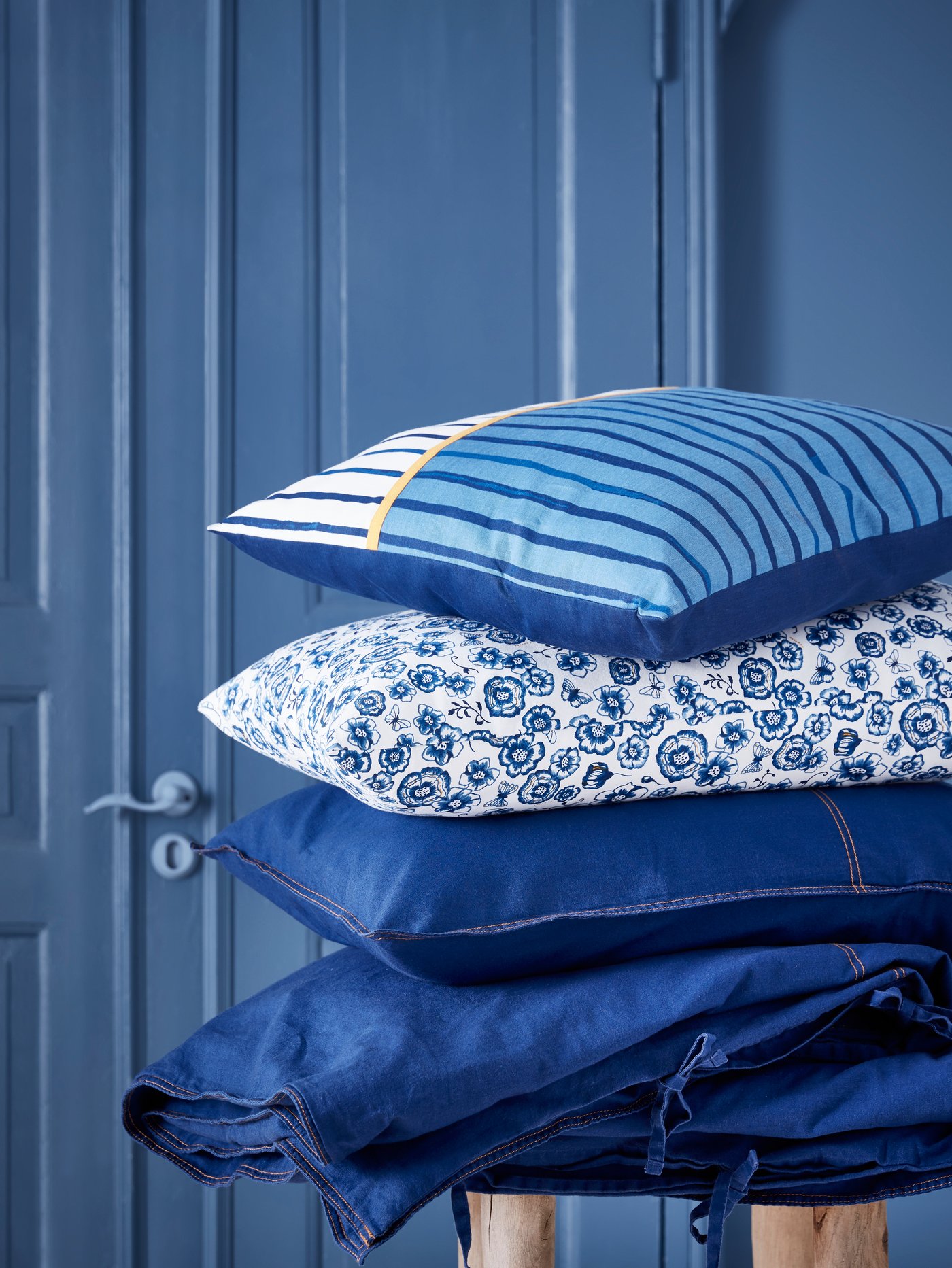 Du linge de lit SÅNGLÄRKA et des coussins SÅNGLÄRKA ornés de différents motifs sont empilés dans une pièce bleue.