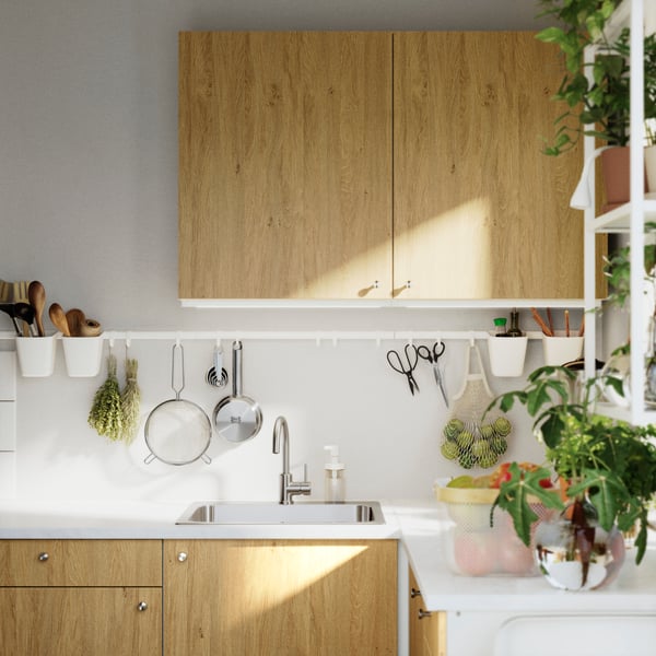 A close-up of a KNOXHULT wall cabinet with doors in a light wood finish, set in a modern kitchen with utensils and plants.