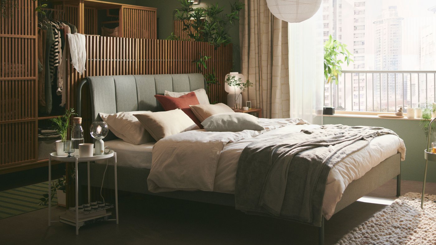A grey-green TÄLLÅSEN upholstered bed dressed with light grey-beige ÄNGSLILJA bed linen, plus cushions and a bedspread.
