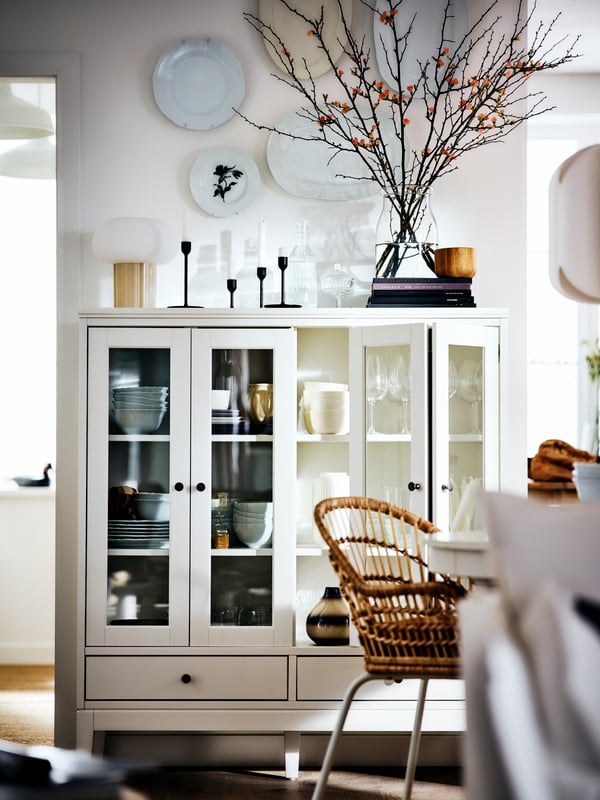 White IDANAS sideboard with glass doors next to a white wall with hanging dishes. Inside, there are several serving dishes and on top of it a pitcher with dry branches, candelabra and a lamp.