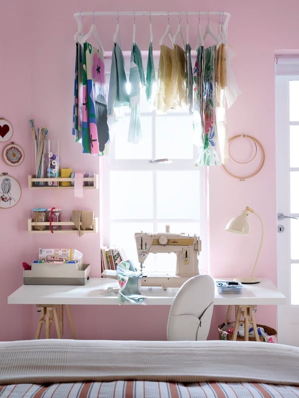 A desk with a sowing machine in front of a pink wall and window with bright light shining through. Textiles hanging above.