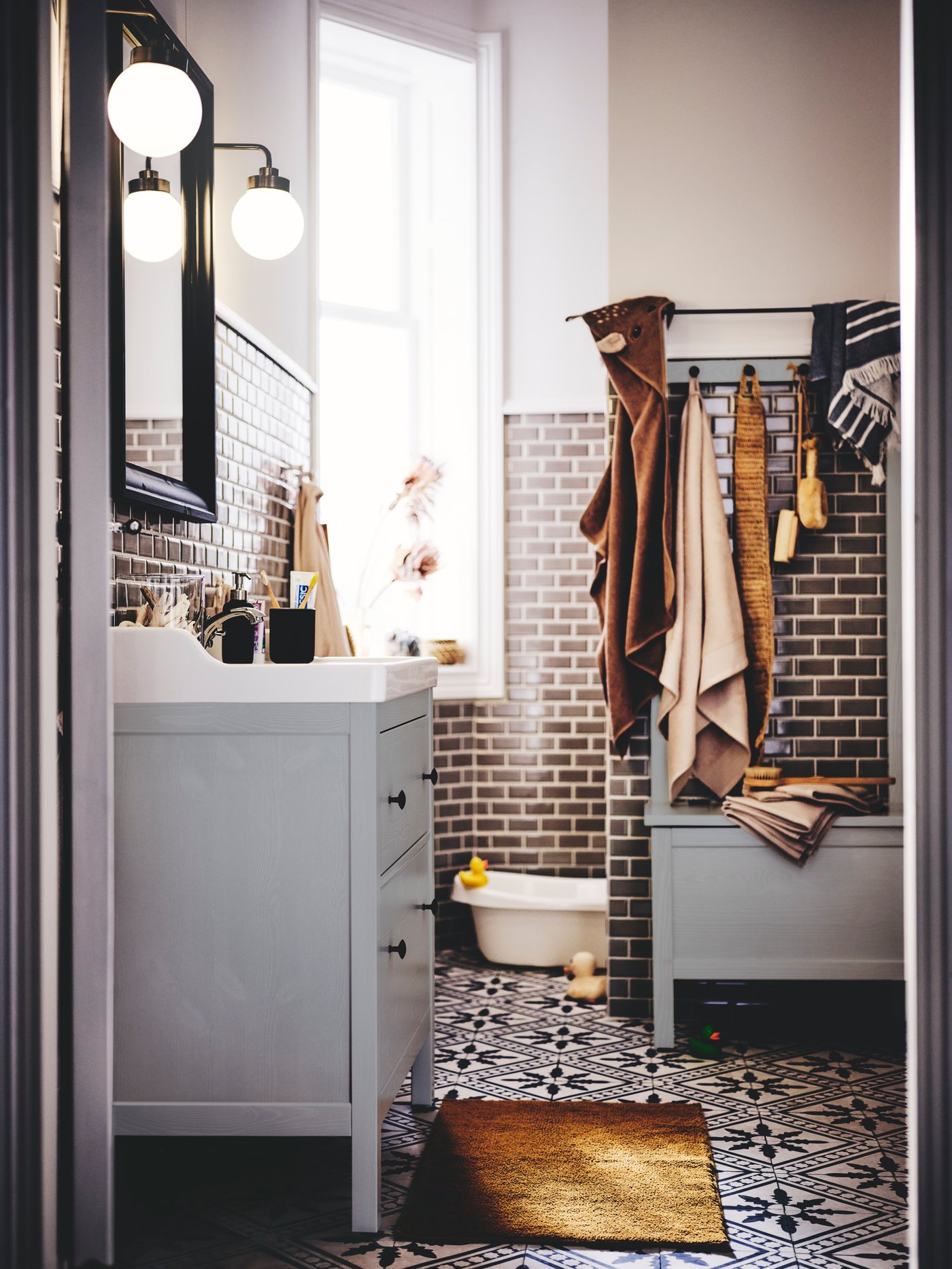 A bathroom with a grey HEMNES/RÄTTVIKEN wash-stand with lamps, a storage bench, a baby bath, bath mat and towels.