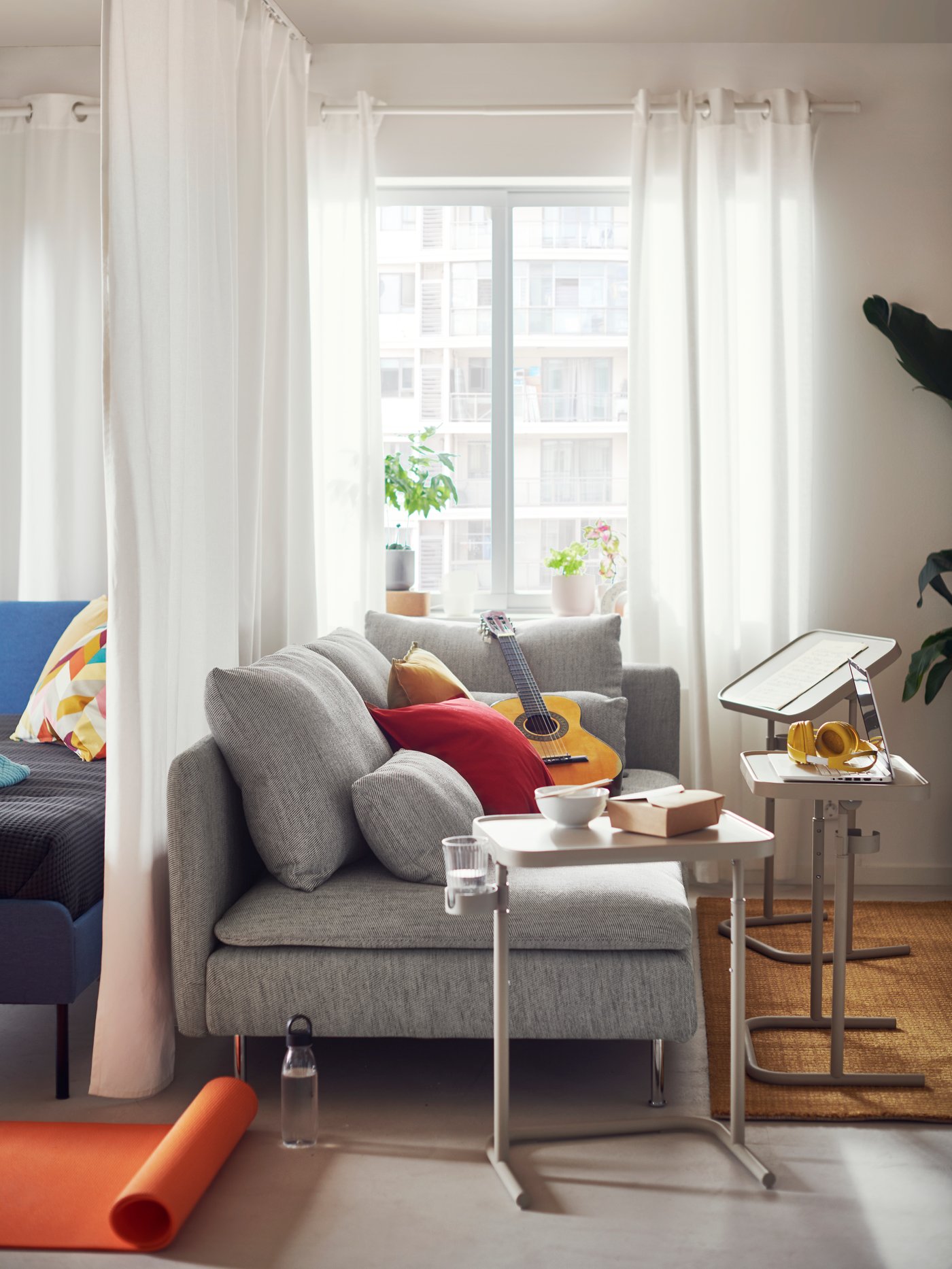 A SÖDERHAMN 3-seat sofa, with a guitar on top of it and three BJÖRKÅSEN laptop stands beside it, stands in front of a window.