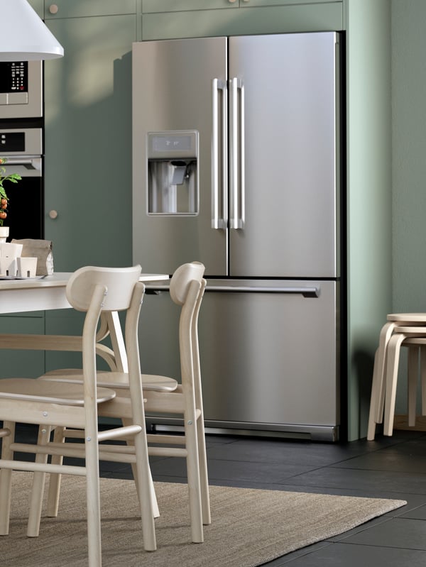 A light-green kitchen with a STJÄRNSTATUS refrigerator in stainless steel. Two pale wooden chairs stand on a rug in front.