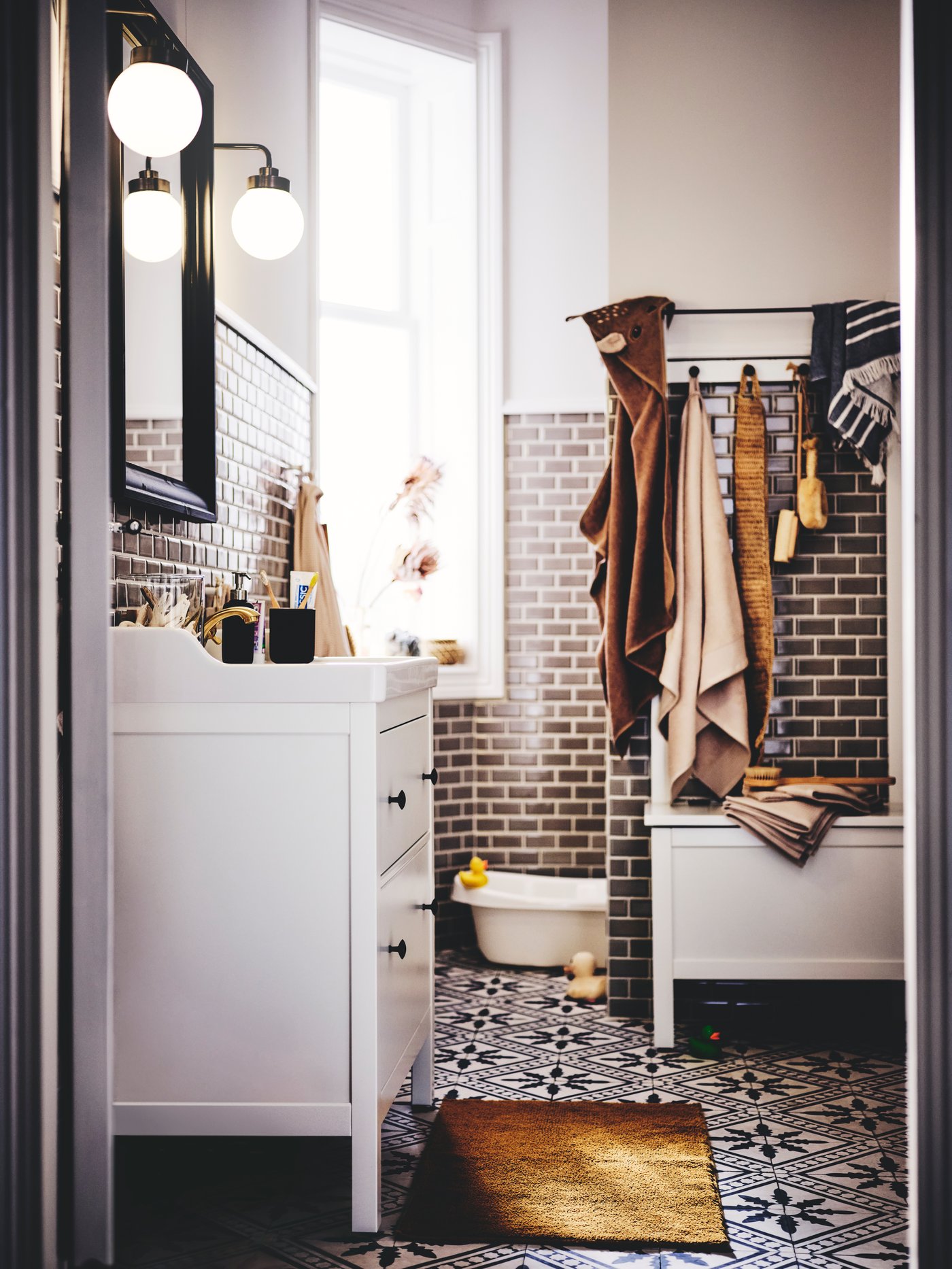 حمام به وحدة حوض غسل HEMNES/RÄTTVIKEN بيضاء، ومصابيح حائط ومقعد تخزين وحوض استحمام للأطفال ولبادة حمام ومناشف.