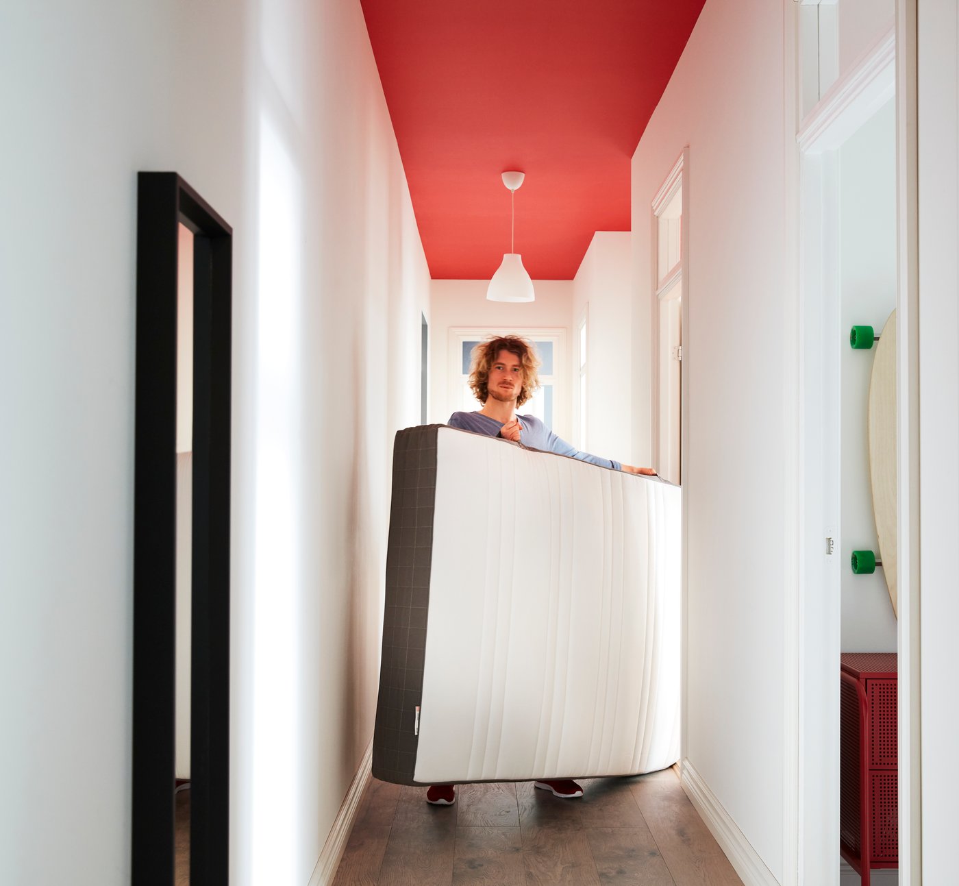 A person carries an upright mattress through a narrow hallway with white walls and a red-painted ceiling.
