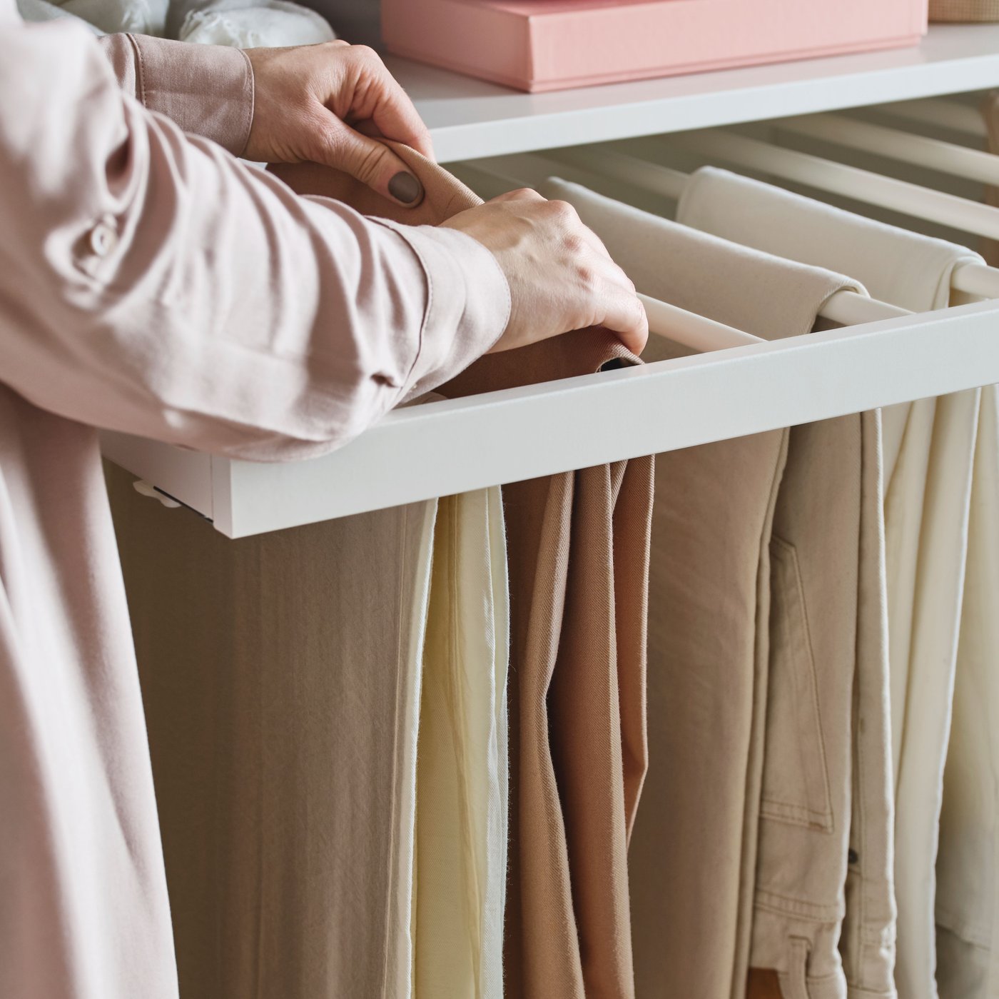 Une personne vêtue d'une chemise rose ajuste un pantalon beige, l'un des nombreux pantalons suspendus à un porte-pantalons coulissant blanc de la marque KOMPLEMENT.