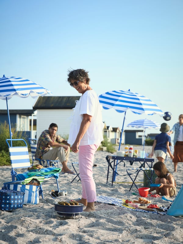 Une famille pique-niquant à la plage, avec des chaises, une table et des tabourets STRANDÖN. La grand-mère gère la cuisson sur le gril STRANDÖN.