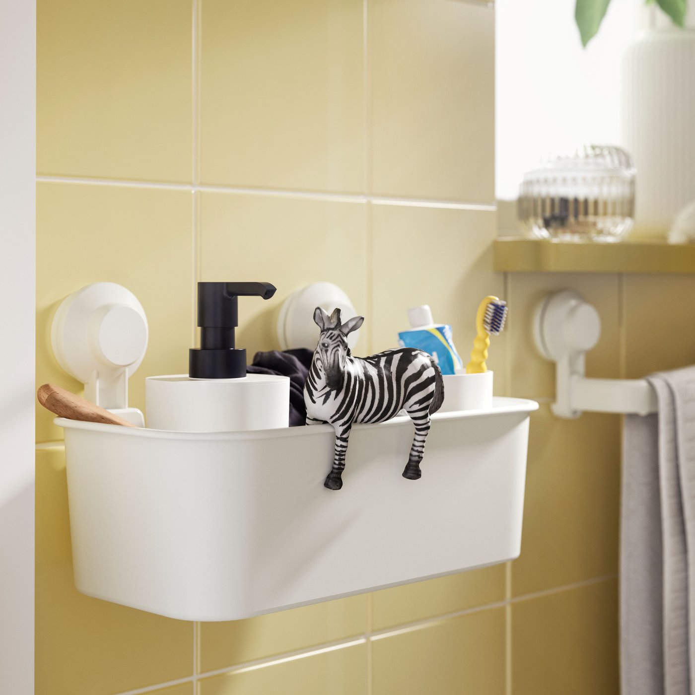 A white plastic TISKEN basket with suction cups, holding toiletries and a toy zebra, is on a yellow-tiled bathroom wall.