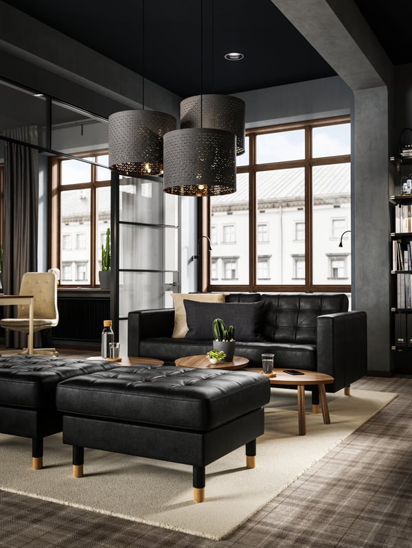 A lounge area created with a dark LANDSKRONA sofa and footstools with small wooden tables and three hanging lamps.
