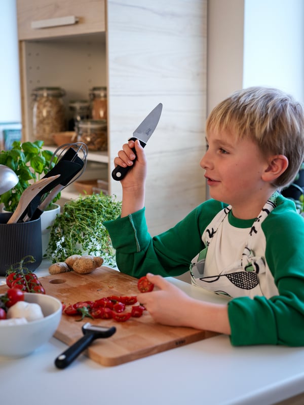 Boy holds a black SMÅBIT knife up as he works at a PROPPMÄTT chopping board with sliced tomatoes and SMÅBIT peeler.