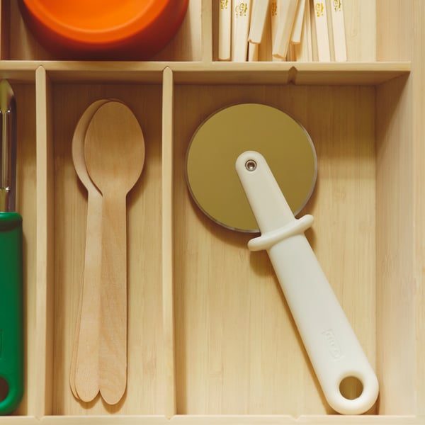 An open drawer with an UPPDATERA cutlery tray in light bamboo holding an UPPFYLLD pizza cutter in off-white and other items.