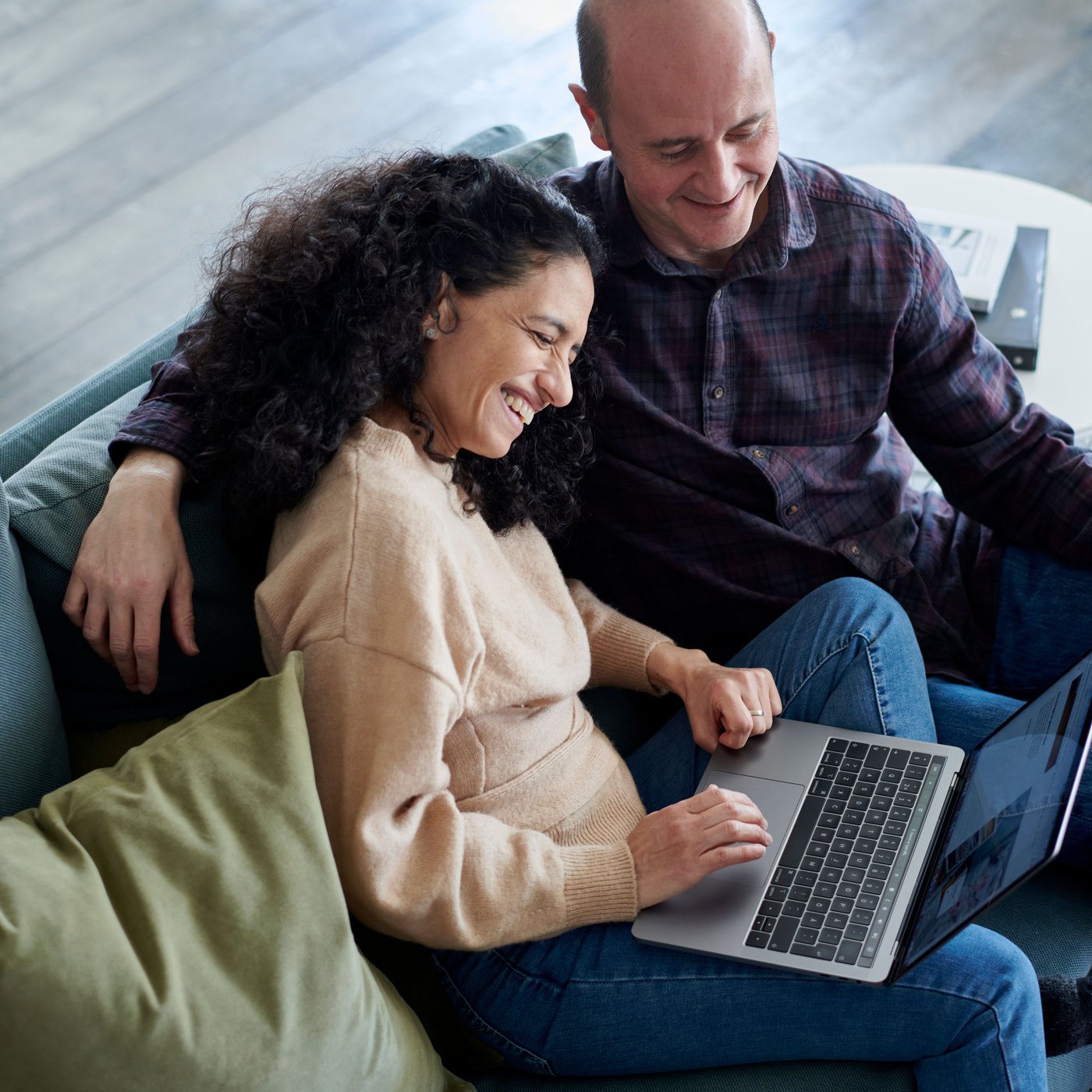 Two people are sitting together on a sofa with a laptop on their laps. One person has curly hair and is wearing a beige sweater, while the other is wearing a dark checkered shirt. Green cushions and the sofa's armrests are visible in the foreground.