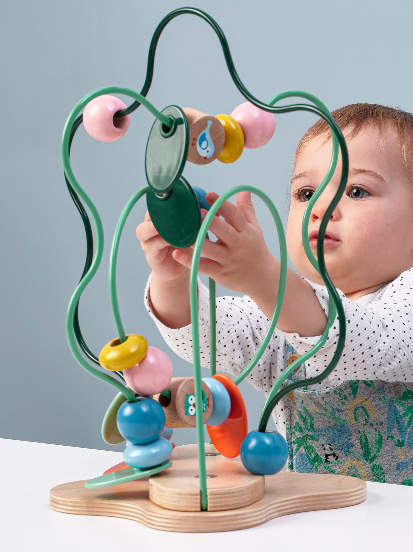 Un bébé vêtu d’un chandail blanc à pois noirs est assis à une table blanche. Il joue avec un colimaçon UPPSTÅ multicolore.