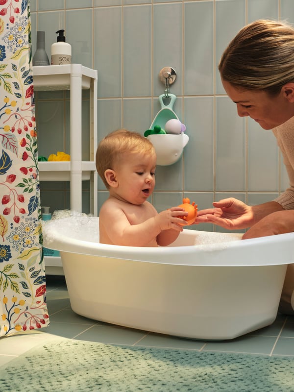 A bathroom where a baby is sitting in a white LÄTTSAM baby bath, with the mother sitting next to it to help.