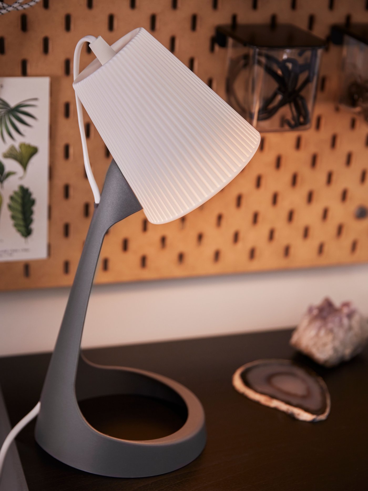 A lit white and dark grey SVALLET work lamp on a desk and a pegboard with a container with lid on the wall behind.