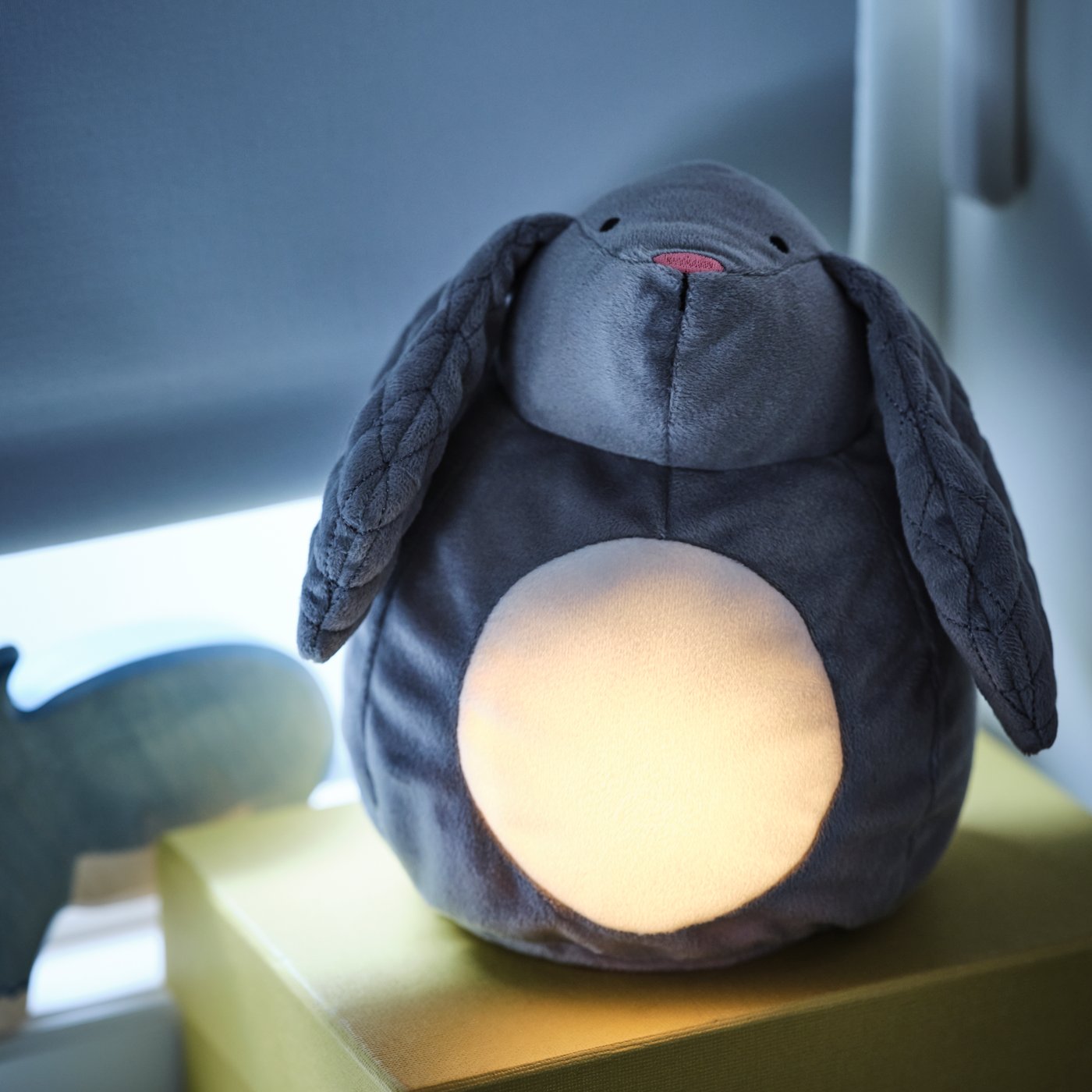Un peluche con luz PEKHULT en forma de conejo azul grisáceo, apoyado sobre una caja amarilla frente a una ventana con la cortina cerrada. 