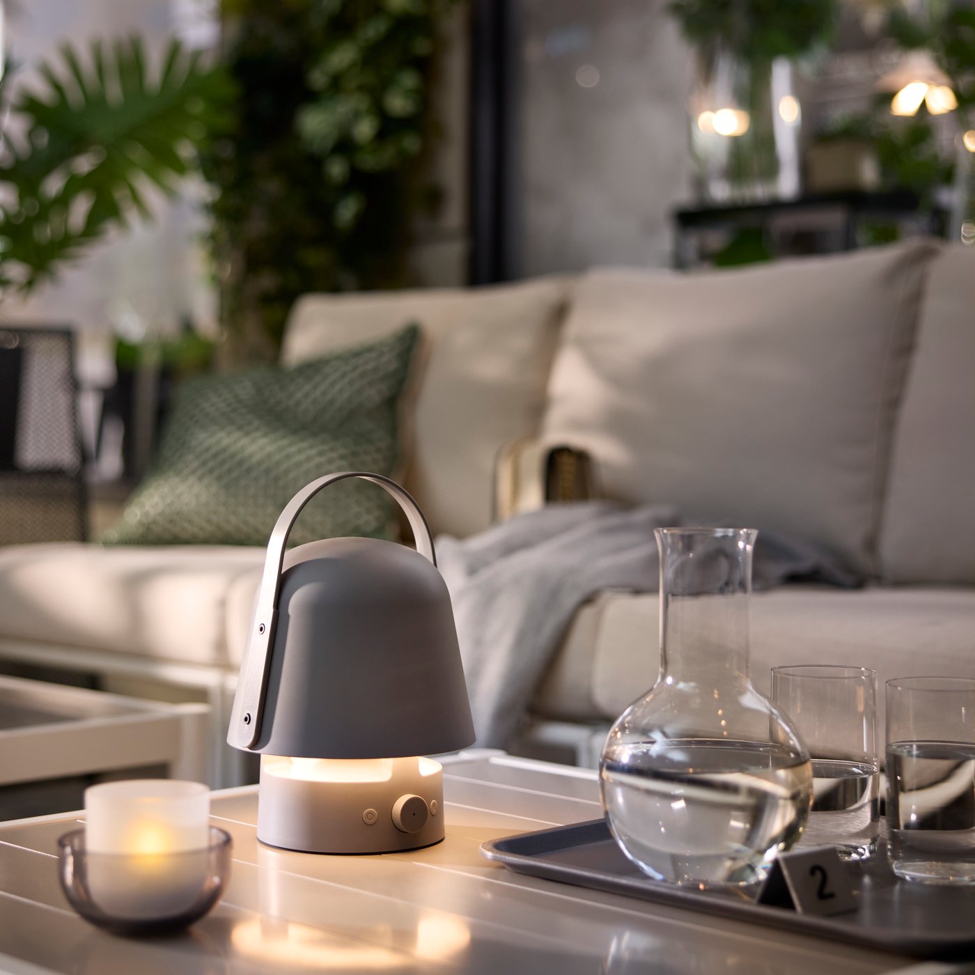 Siva VAPPEBY bluetooth zvučnik-lampa na stolu okružena biljkama, čašama i svećama u bašti kafića.