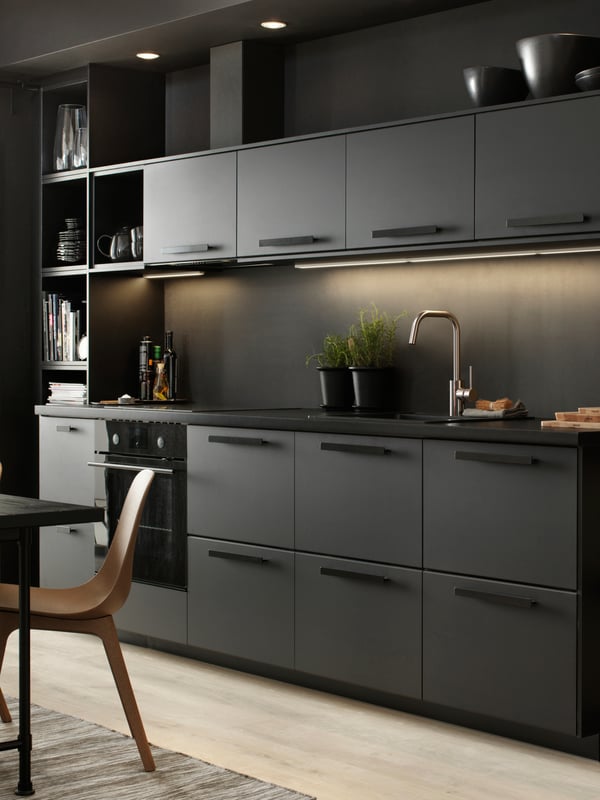 A sleek black kitchen with lighting under the top cupboards. Two black pots with plants inside stand on the counter.