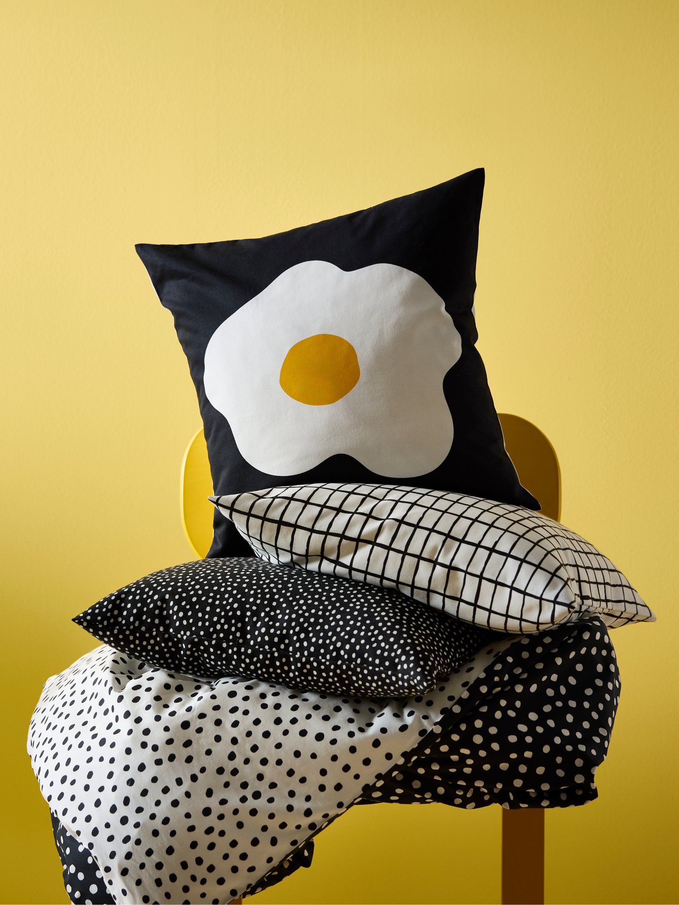 Bold black and white patterned cushions piled on top of a yellow chair. An egg-themed cushion rests on top.
