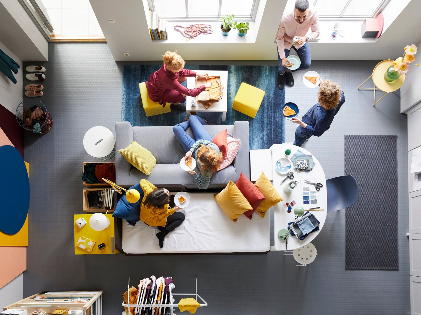 View from above of a living room where several people are sitting and standing around a sofa and small tables, eating and socializing with food and drinks in front.