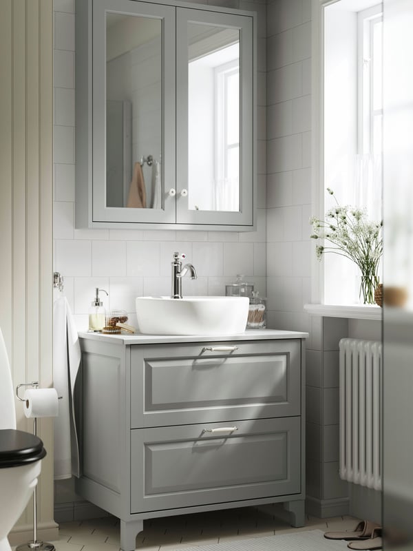 A white-tiled bathroom has a light-grey TÄNNFORSEN vanity with two drawers and a mirror cabinet with two doors above it.