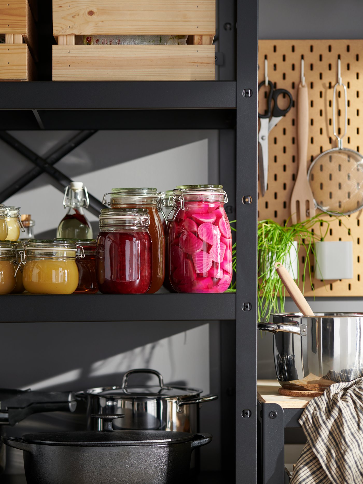 Köksförvaring med hylla fylld med trälådor, inläggningar i KORKEN burkar, kastruller samt köksredskap på en pegboard och en krukväxt bredvid.