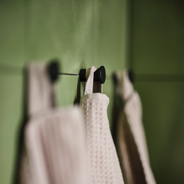 Dark beige towels hanging from three black SKOGSVIKEN hooks against a green tiled wall.