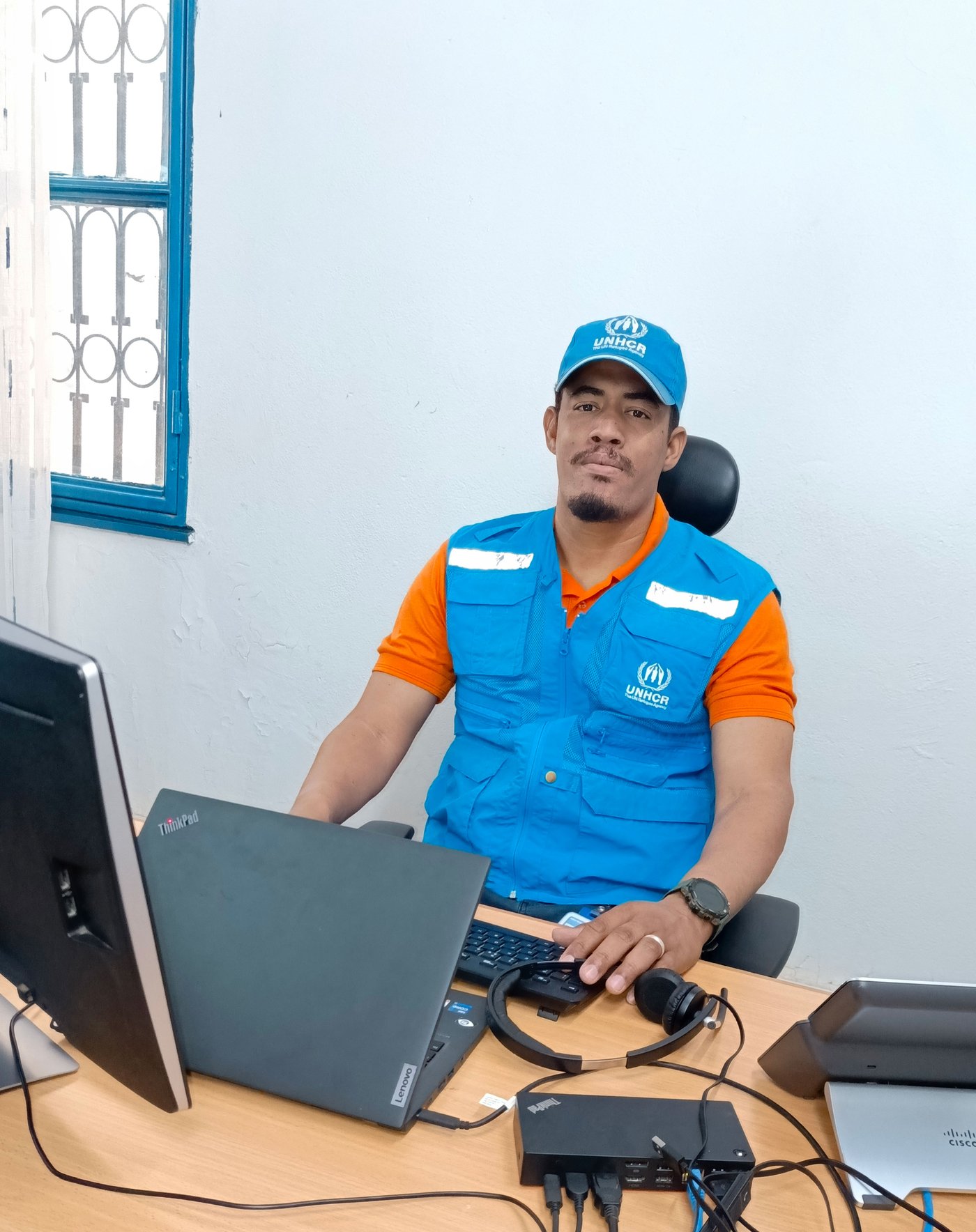 Dans son uniforme bleu du HCR, Ilyas assis devant un ordinateur dans un bureau.
