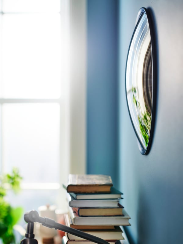 A black SVARTBJÖRK decorative convex mirror hangs on a blue wall above a pile of books near a window in a living room.