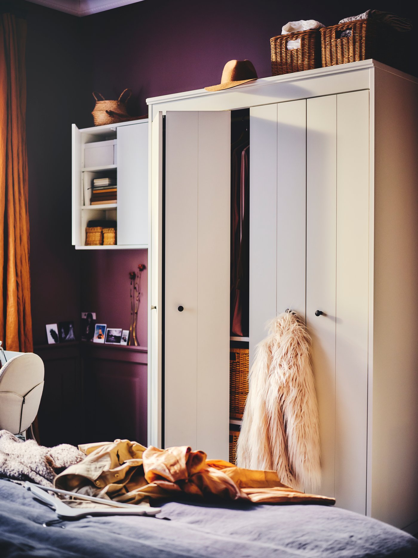 A white IDANÄS wardrobe with one open folding door and a jacket hanging from a knob stands against a wall near a bed.