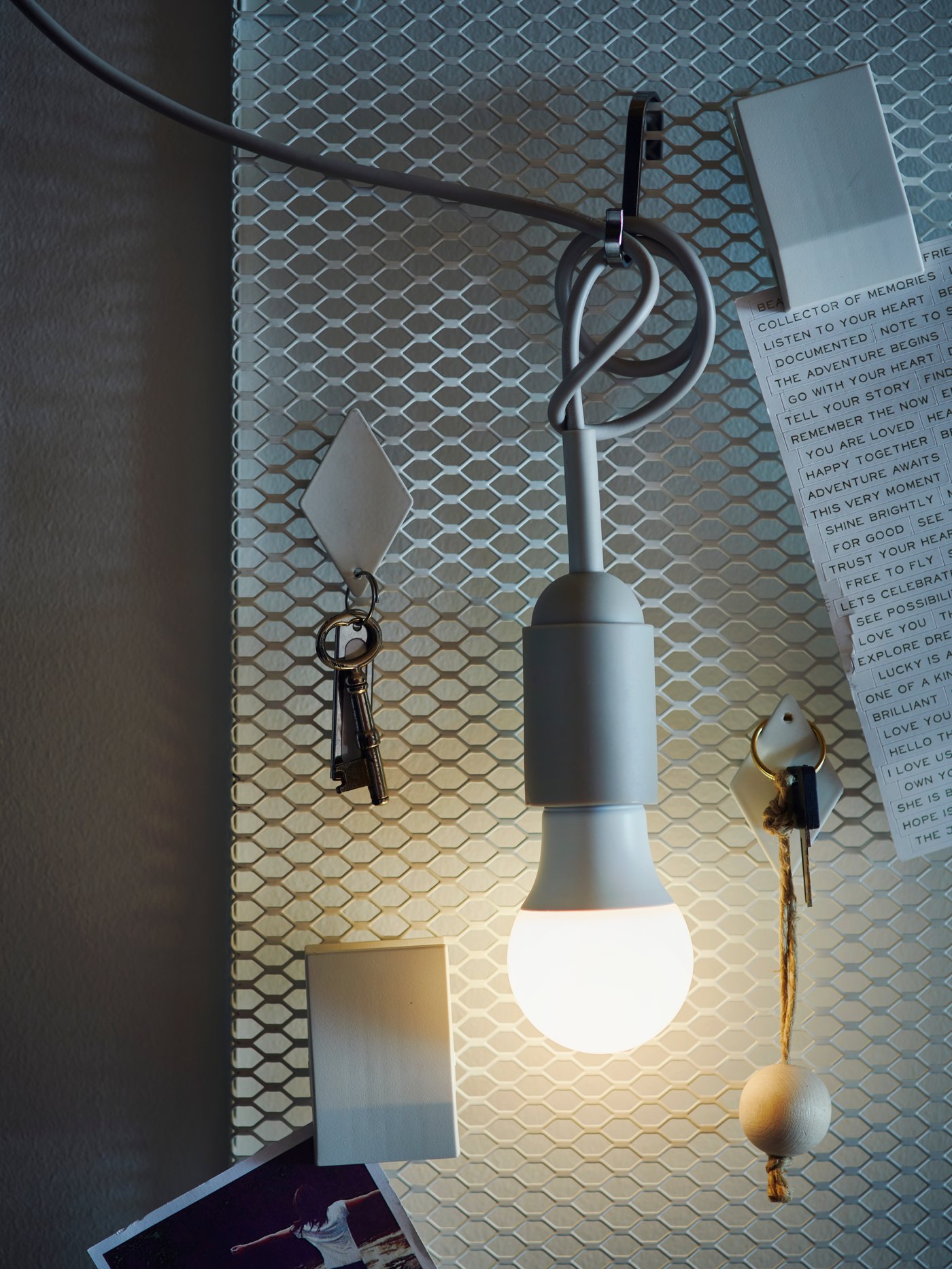 A lit SOLHETTA LED bulb is hanging from a hook that is attached to a wire mesh wall that also holds keys and notes.