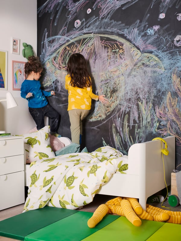 Two children stand on a white BUSUNGE bed dressed in a BLÅVINGAD green turtle duvet cover and draw on a chalkboard wall.