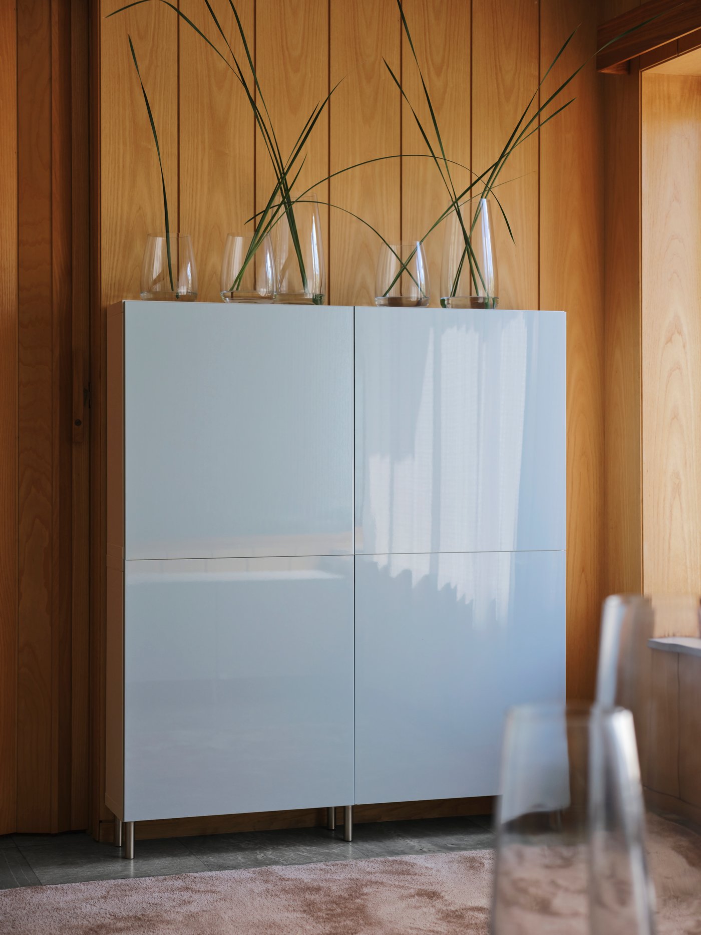 A BESTÅ frame standing against a wooden panelled wall has four SELSVIKEN doors and glass vases sitting on top.