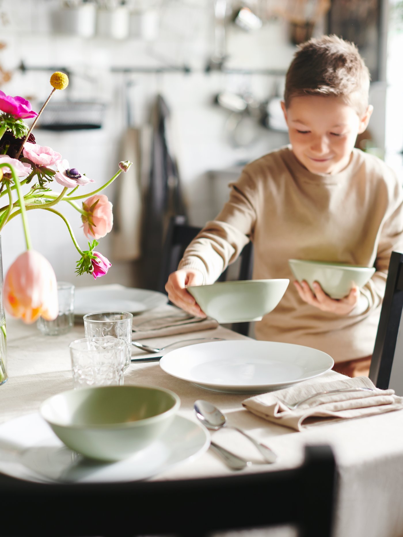 A sunlit table with a tablecloth and flowers is being set by a boy distributing matt-green FÄRGKLAR bowls onto white plates.